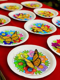 paper plates with butterflies painted on them are sitting on a red tablecloth covered table