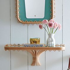 a mirror and vase with flowers on a shelf in front of a white painted wall