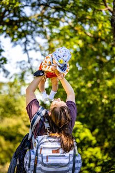 a woman holding a baby up in the air with her hands above her head and trees behind her