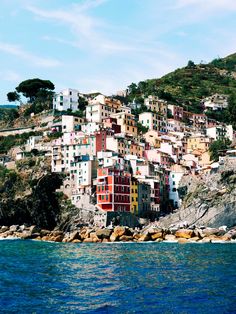 colorful buildings on the side of a hill next to blue water and rocks in front of it