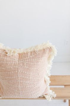 a pink pillow with fringe on it sitting on a wooden bench in front of a white wall