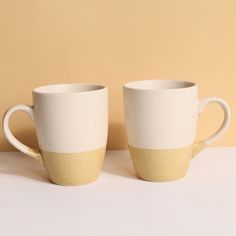 two white and yellow coffee mugs sitting next to each other on a beige background