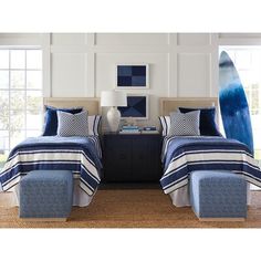 two beds with blue and white bedding in a room next to windows, one has a surfboard on the wall