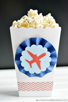 an airplane themed popcorn box with blue and red ribbon around the top, sitting on a white table