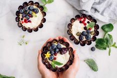 two hands holding small desserts with berries and blueberries in them on a marble surface