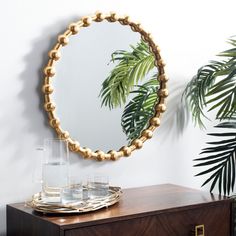 a round mirror sitting on top of a wooden dresser next to a plant and glasses