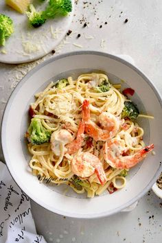 a white bowl filled with pasta and shrimp on top of a table next to broccoli