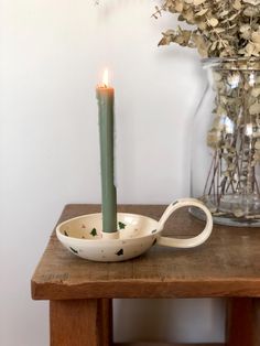 a lit candle sits in a bowl on a table next to a vase with dried flowers