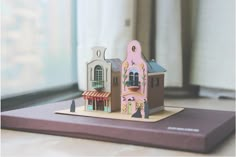 a small wooden toy house on top of a table next to a window with curtains