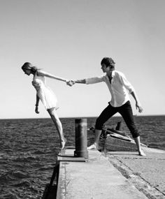 a man and woman holding hands while standing on a pier near the ocean, black and white photograph