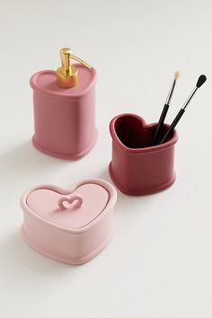 two pink heart shaped containers with makeup brushes in them on a white surface next to each other