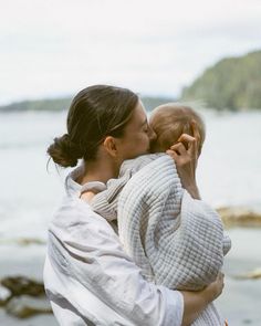 a woman holding a baby in her arms by the water's edge while kissing it