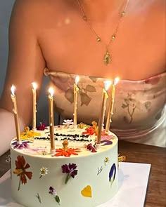 a woman sitting in front of a birthday cake with lit candles on it and flowers