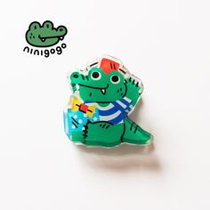 a green and red alligator pin sitting on top of a white table next to a sticker