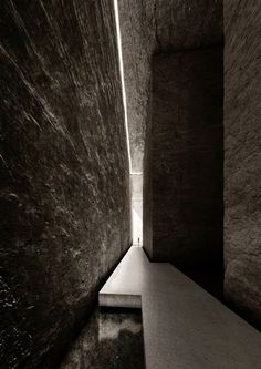 a long narrow hallway with concrete walls and water running down the side wall, leading to another room