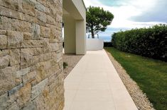 a walkway between two buildings with grass and shrubs in the background, along side it is a stone wall