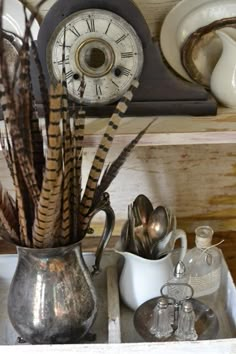 an old clock is sitting on the shelf next to dishes and utensils in a silver pitcher