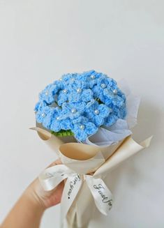 a hand holding a bouquet of blue crocheted flowers with white ribbon around it