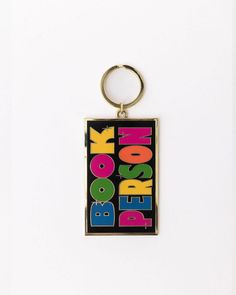 enamel keychain with black rectangle with colorful 'book person' across the front Reading Supplies, Enamel Keychain, Calendar Notebook, Tech Pouch, Tunnel Of Love, Desk Planners, Host Gifts, Pill Case, Cozy Gift