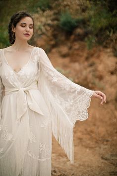 a woman wearing a white dress with long sleeves and fringes on her arms, standing in the dirt