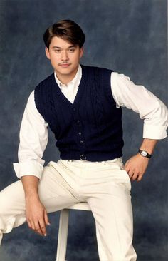 a man sitting on top of a stool wearing white pants and a blue sweater vest