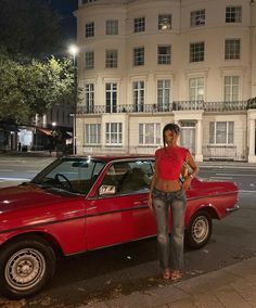 a woman standing next to a red car in front of a white building at night