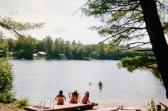 Image is of a lake. There are a few people sitting by the lake on beached docks and a few people in the water. Camp Lake Aesthetic, 1980s Summer Camp Aesthetic, Vintage Summer Camp Aesthetic, 80s Summer Camp Aesthetic, American Summer Aesthetic, Lake Summer Aesthetic, Vintage Summer Camp, American Summer Camp, Slasher Summer