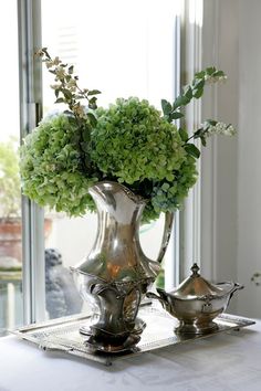 a silver vase filled with green flowers sitting on top of a table next to a window