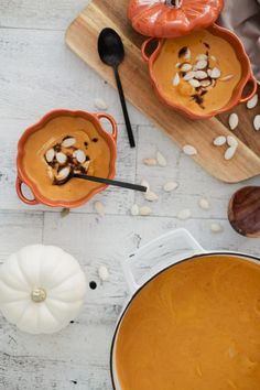 two bowls of pumpkin soup on a cutting board with spoons and other ingredients around them