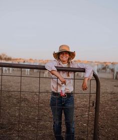 a woman wearing a cowboy hat leaning on a fence with cattle in the background at sunset