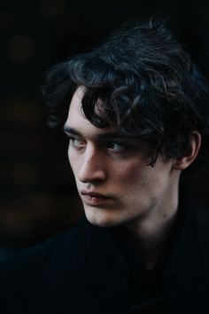 a young man with curly hair looks into the distance while wearing a black shirt and tie