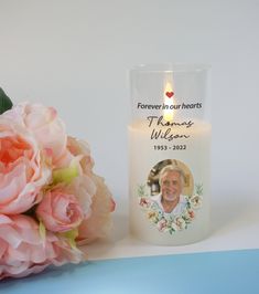 a white candle with a photo on it next to pink flowers and a blue background