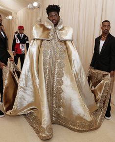 two men standing next to each other in front of a white wall wearing gold and silver gowns