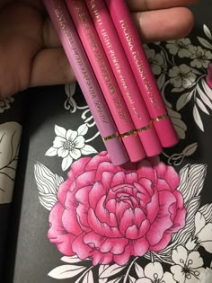 three pink pencils are held in front of a floral wallpapered surface with flowers on it