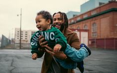 a man holding a child in his arms and smiling at the camera with buildings behind him