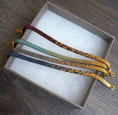 three bracelets in a box on a wooden table, one is gold and the other two are multicolored