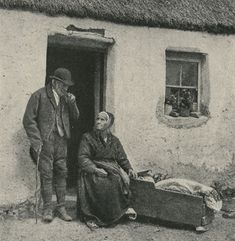 an old black and white photo of two people standing in front of a thatched house