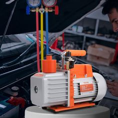 a man looking at an orange and white device on top of a car engine with other vehicles in the background