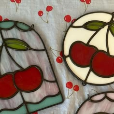 two stained glass pieces with cherries and leaves on a white tablecloth, one is red