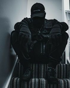 a man sitting on top of a stair case wearing a black jacket and hat with his hands behind his head