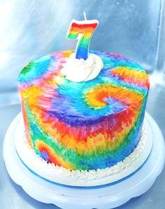 A cake is painted with rainbow tie dye and has a number 7 candle on the top. It's on a turntable on a stainless steel counter Tie Dye Smash Cake, Tie Dye Cake Frosting, Tie Dye Cake Pops