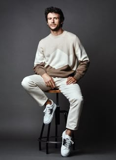 a man sitting on top of a stool wearing white pants and a brown sweater,