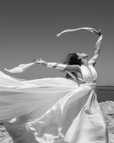 a woman in a long white dress is standing on rocks and holding her arms out