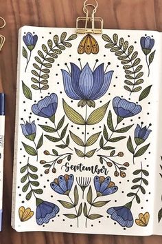 an open notebook with blue flowers on it next to some markers and pens, along with a clipboard