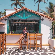 a woman sitting at a table in front of a bar with palm trees behind her