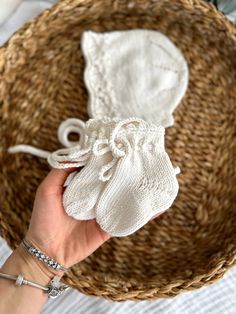 a hand holding a white knitted baby bonnet in a wicker basket on top of a bed