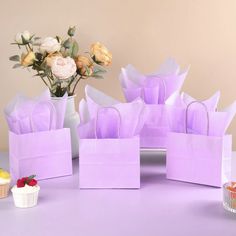 purple paper bags sitting on top of a table next to cupcakes and flowers