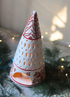 a white christmas tree shaped candle holder with red and white designs on the top, surrounded by evergreen branches