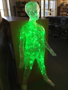 a green light up man standing in front of a book shelf with chairs behind it