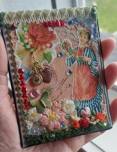 a person holding up a small square piece of art with flowers and charms on it